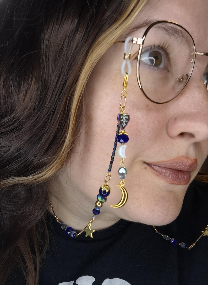 May include: A gold-toned eyeglass chain featuring a variety of blue beads, iridescent heart-shaped charms, a crescent moon charm, and a small star charm. The chain attaches to the frames with white silicone loops and gold-toned metal connectors.