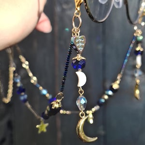 May include: A gold-toned eyeglass chain featuring dark blue beads, iridescent heart-shaped charms, white crescent moon beads, and metallic gold crescent moon pendants. The chain attaches to glasses with gold lobster clasps and clear silicone loops.