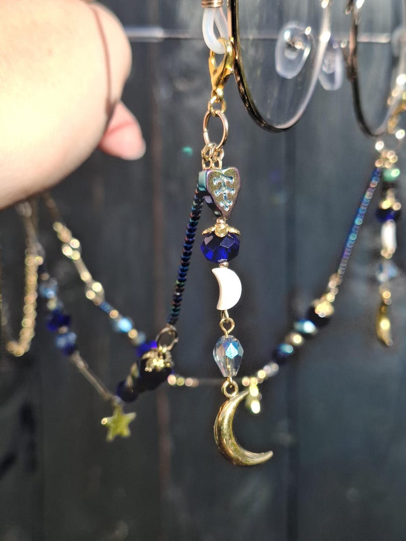 May include: A gold-toned eyeglass chain featuring dark blue beads, iridescent heart-shaped charms, white crescent moon beads, and metallic gold crescent moon pendants. The chain attaches to glasses with gold lobster clasps and clear silicone loops.