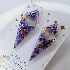 Könnte beinhalten: Ein Paar handgefertigte, lila Harzohrringe mit Goldflocken und schillerndem Glitzer. Die Ohrringe sind dreieckig mit einem kreisförmigen Ausschnitt und goldfarbenen Ringen zur Befestigung. Der Text "SPACE JOURNEY" ist sichtbar.