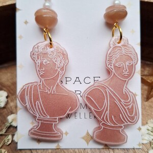 May include: A pair of dangle earrings featuring a bust of a classical figure. The earrings have gold-colored hoops, a peach-colored bead, and a pearl. The bust is peach-colored with white outlines. The earrings are displayed on a white card with gold stars.