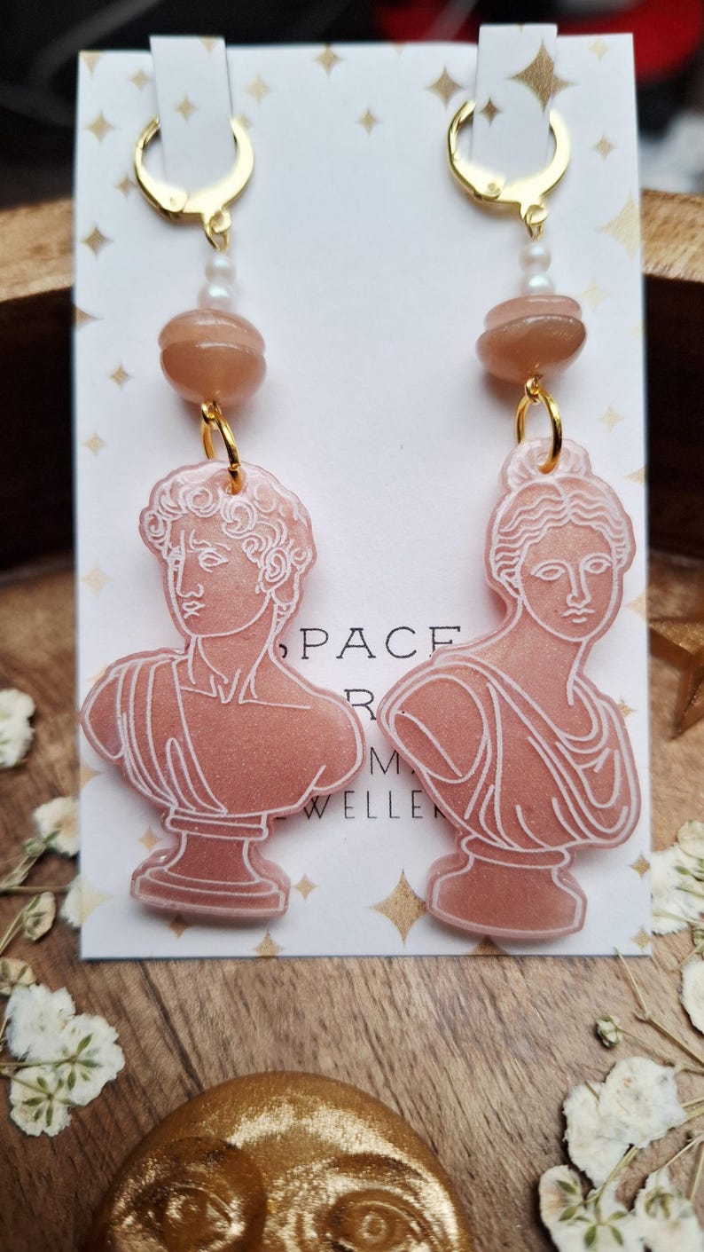 May include: A pair of dangle earrings featuring a bust of a classical figure. The earrings have gold-colored hoops, a peach-colored bead, and a pearl. The bust is peach-colored with white outlines. The earrings are displayed on a white card with gold stars.