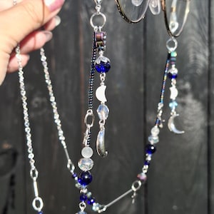 May include: A silver-toned eyeglass chain featuring dark blue beads, iridescent clear beads, and small silver crescent moon charms. The chain attaches to the frames with clear rubber loops and includes metallic links and dark seed beads.