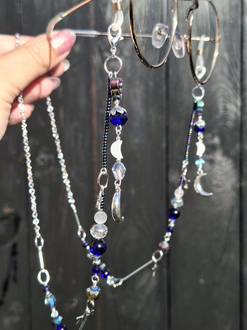 May include: A silver-toned eyeglass chain featuring dark blue beads, iridescent clear beads, and small silver crescent moon charms. The chain attaches to the frames with clear rubber loops and includes metallic links and dark seed beads.