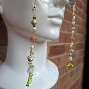 May include: Eyeglass chain with gold-coloured hardware and a variety of beads. The chain features clear, iridescent, and pearl-like beads, along with small green leaf charms. The chain is attached to a pair of eyeglasses.