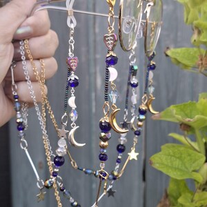 May include: A pair of gold-tone metal eyeglasses featuring decorative beaded chains attached to the temples. The chains are adorned with dark blue beads, iridescent crystals, silver-tone star charms, and crescent moon pendants in gold and silver tones.