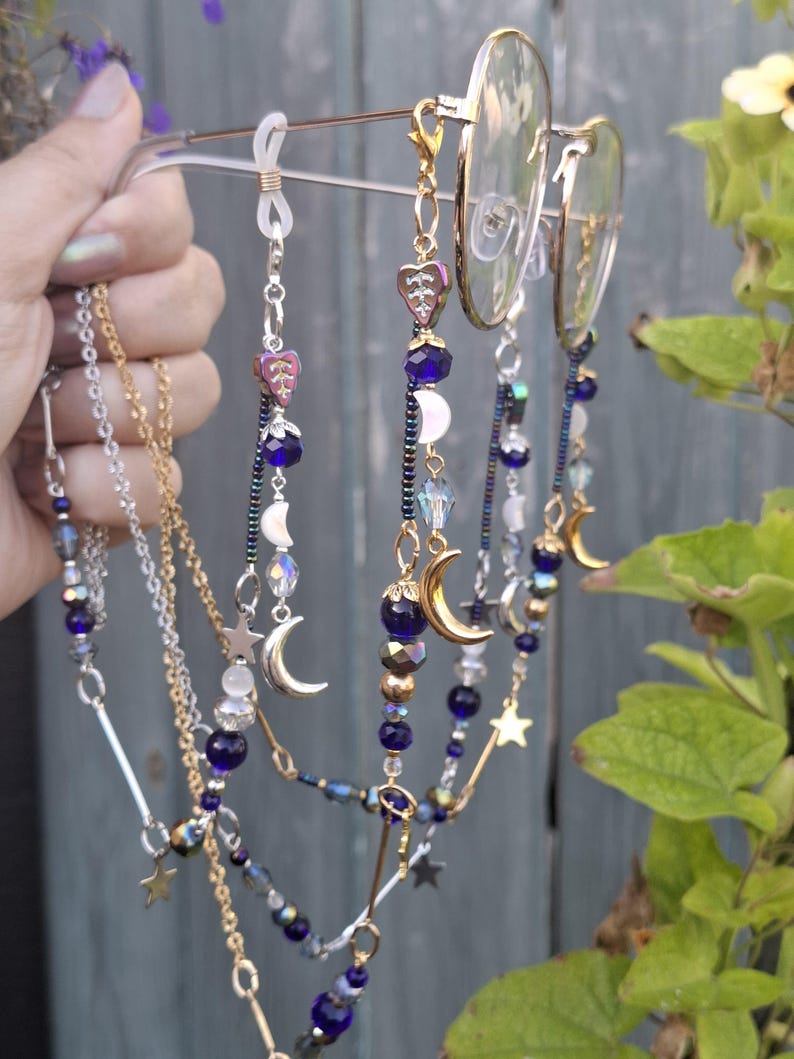 May include: A pair of gold-tone metal eyeglasses featuring decorative beaded chains attached to the temples. The chains are adorned with dark blue beads, iridescent crystals, silver-tone star charms, and crescent moon pendants in gold and silver tones.