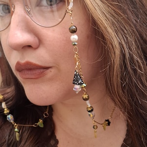May include: A gold-toned eyeglass chain featuring a variety of beads including white pearls, dark metallic spheres, and faceted crystals. The chain includes a small black mushroom charm with white dots and gold-toned metal links connecting the beads.