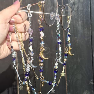 May include: A pair of gold-tone metal eyeglasses featuring multiple decorative beaded chains attached to the frames. The chains include dark blue, clear, and iridescent beads, along with gold and silver-tone metal charms shaped like crescent moons and stars.