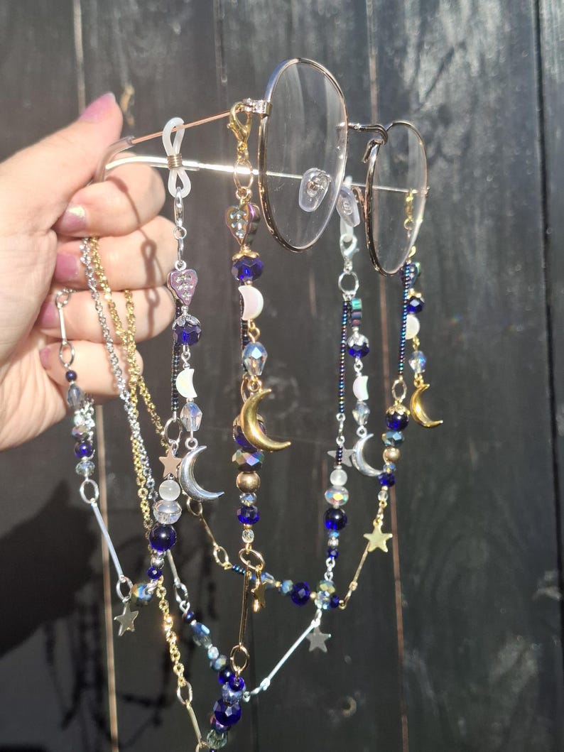 May include: A pair of gold-tone metal eyeglasses featuring multiple decorative beaded chains attached to the frames. The chains include dark blue, clear, and iridescent beads, along with gold and silver-tone metal charms shaped like crescent moons and stars.