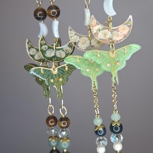 May include: Dangling earrings featuring gold-tone chains, crescent moon charms with floral patterns, and green moth pendants. The designs include tiger eye, clear crystal, and pearl-like beads, suspended from gold-tone lobster claw clasps.