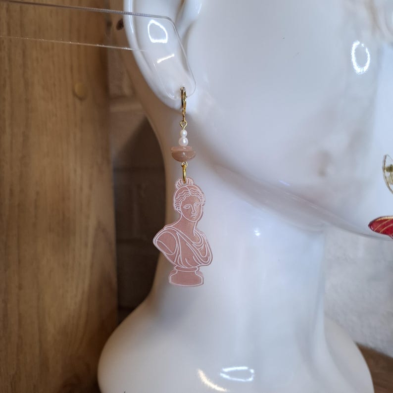 May include: A pair of dangle earrings featuring a pink bust of a classical figure. The earrings have gold-colored hooks and are adorned with small pearls and a pink stone. The earrings are displayed on a white mannequin head.
