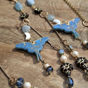 May include: A pair of gold-tone chain necklaces featuring light blue moth pendants with gold detailing, white crescent moon charms, light blue oval beads, black flower-shaped beads, white round beads, and dark gray faceted crystal beads.