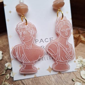 May include: A pair of dangle earrings featuring pink resin busts of classical figures. Each earring has a gold-colored hoop, a string of small pearls, and a peach-colored bead. The earrings are displayed on a white card with gold star accents.