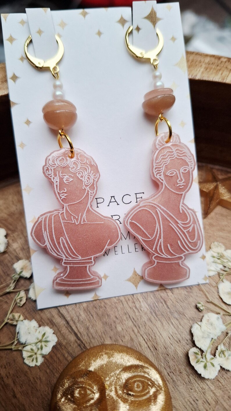 May include: A pair of dangle earrings featuring pink resin busts of classical figures. Each earring has a gold-colored hoop, a string of small pearls, and a peach-colored bead. The earrings are displayed on a white card with gold star accents.