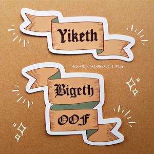 Puede incluir: Dos pegatinas decorativas con un diseño de banner. La pegatina superior presenta la palabra "Yiketh" en una fuente gótica negra sobre una pancarta de color canela. La pegatina inferior presenta las palabras "Bigeth" y "OOF" en una fuente gótica negra sobre una pancarta de color canela y verde.