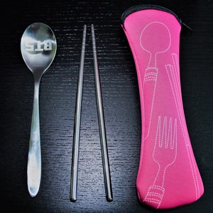 Peut inclure: Un étui de voyage rose avec un contour blanc d'une cuillère, d'une fourchette et de baguettes. L'étui contient une cuillère en argent avec le texte "BTS" gravé dessus et deux baguettes en argent.