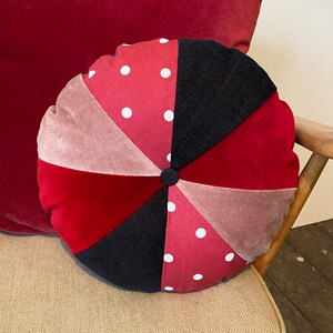 May include: Round red, pink and black patterned throw pillow with white polka dots. The pillow has a button in the centre.
