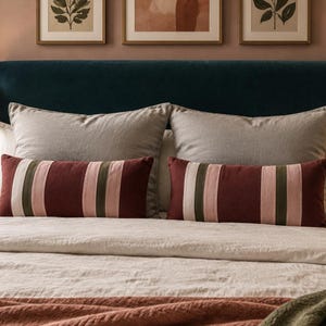 May include: Two rectangular lumbar cushions featuring vertical stripes in shades of deep red, olive green, and cream. These cushions are positioned in front of two large, solid light grey square cushions on a bed with a dark blue velvet headboard.