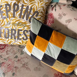 May include: A patchwork cushion with squares of black, mustard yellow, light green, and brown velvet. The cushion is sitting on a floral patterned sofa with a cushion that says "Epping Forest is calling" in grey letters on a yellow background.