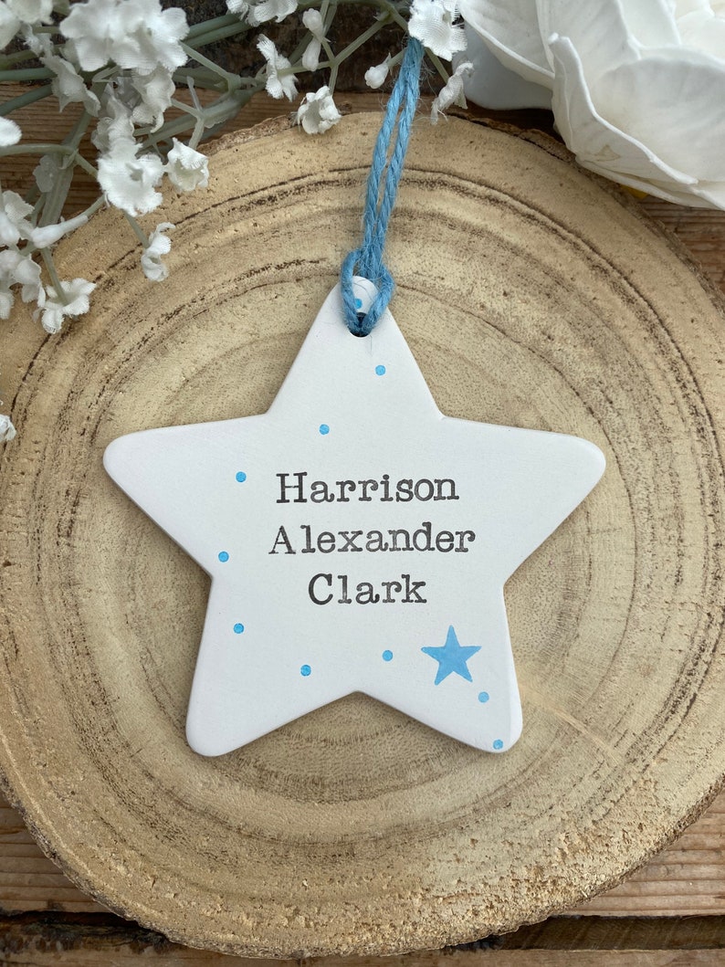 May include: White ceramic star ornament with blue polka dots and a blue star. The ornament has the names "Harrison Alexander Clark" written on it.