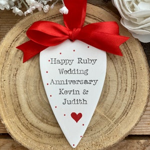 May include: White heart-shaped ceramic ornament with red ribbon and red dots. The ornament says "Happy Ruby Wedding Anniversary Kevin & Judith".