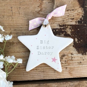 May include: White ceramic star ornament with pink polka dots and the text "Big Sister Darcy".