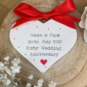 May include: A white heart-shaped ornament with red polka dots and a red bow. The ornament has the text "Nana & Papa 20th July 1985 Ruby Wedding Anniversary" and a red heart. The ornament is displayed on a wooden slice.
