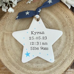May include: White ceramic star ornament with blue dots and a blue star. The ornament has the text "Kyran 25.05.23 12:31 am 5lbs 9ozs" written on it.