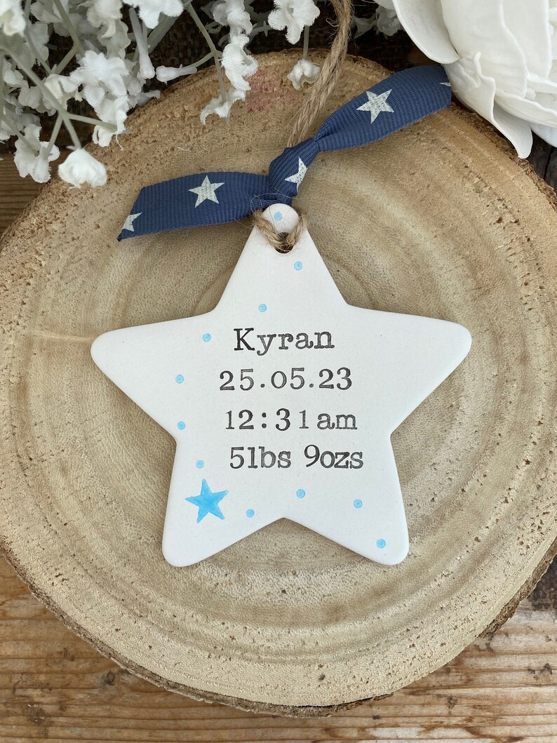 May include: White ceramic star ornament with blue dots and a blue star. The ornament has the text "Kyran 25.05.23 12:31 am 5lbs 9ozs" written on it.