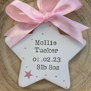 May include: A white ceramic star with pink polka dots and a pink ribbon. The star has the name "Mollie Tucker" and the date "01.02.23" and weight "8lb 8oz" written on it.