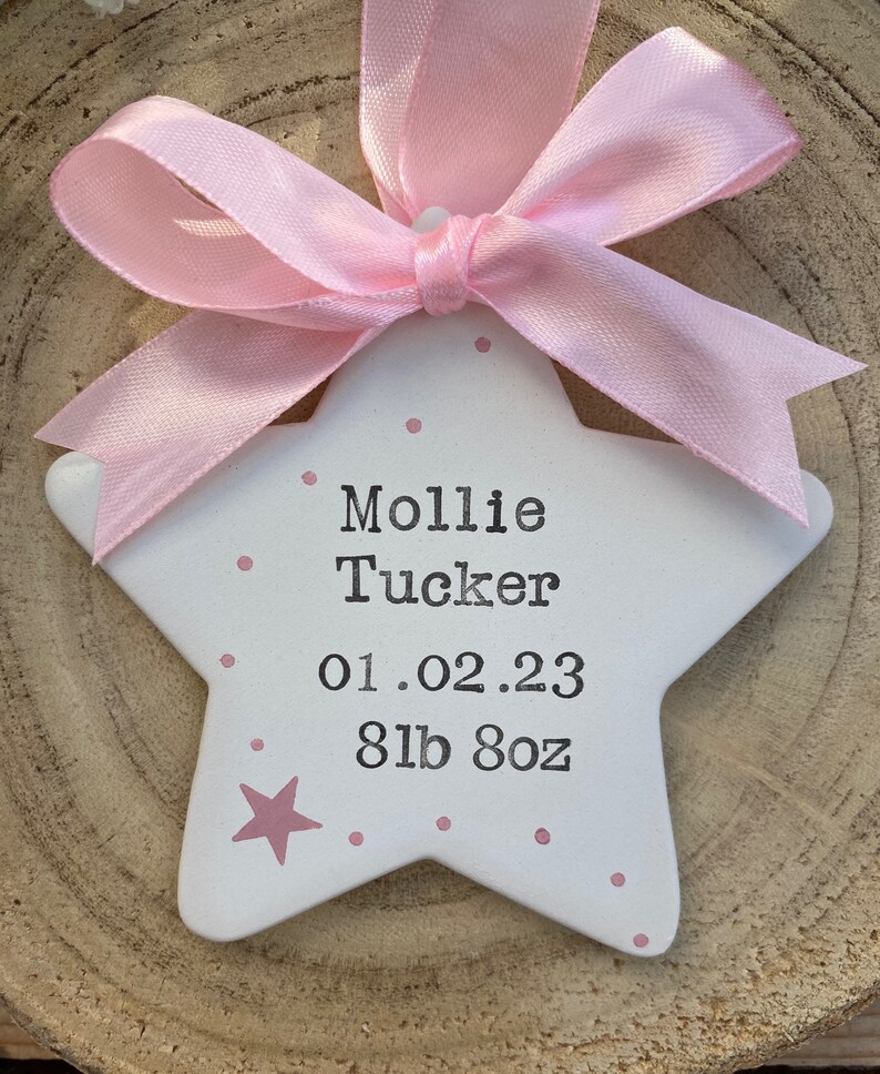 May include: A white ceramic star with pink polka dots and a pink ribbon. The star has the name "Mollie Tucker" and the date "01.02.23" and weight "8lb 8oz" written on it.