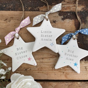 May include: Three white ceramic star ornaments with pink, blue and grey polka dot ribbons. The stars are decorated with pink and blue dots and have the text "Big Sister Chloe", "Little Sister Amaia", and "Big Brother Alex" written on them.