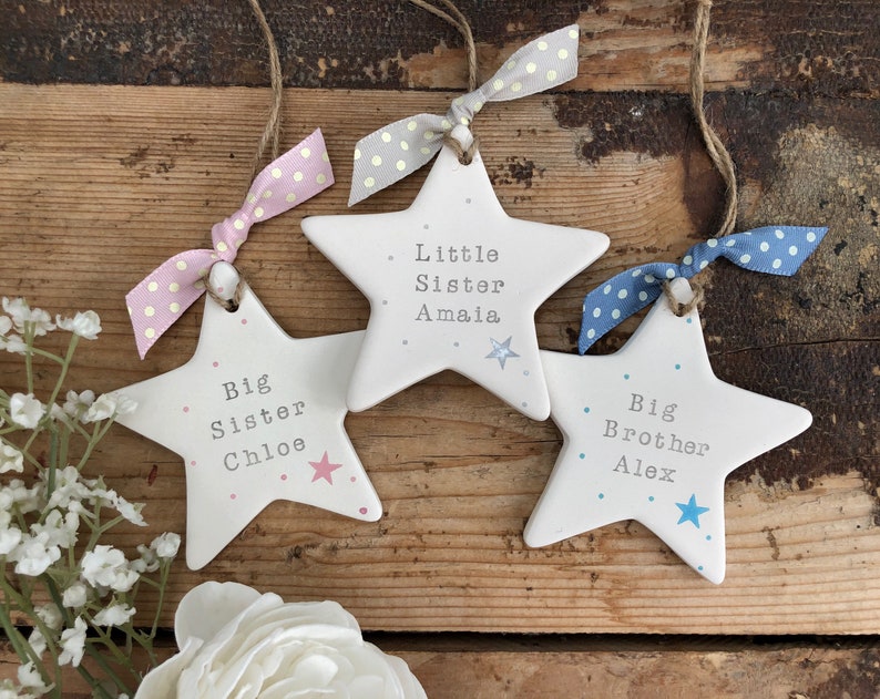 May include: Three white ceramic star ornaments with pink, blue and grey polka dot ribbons. The stars are decorated with pink and blue dots and have the text "Big Sister Chloe", "Little Sister Amaia", and "Big Brother Alex" written on them.
