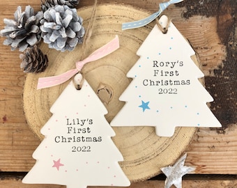 May include: Two white ceramic Christmas tree ornaments with pink and blue accents. The ornaments are decorated with small dots and a star. The text on the ornaments reads "Lilly's First Christmas 2022" and "Rory's First Christmas 2022".