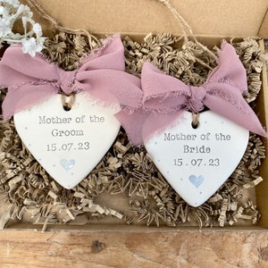 May include: Two heart-shaped ceramic ornaments, one for the Mother of the Groom and one for the Mother of the Bride, with the date 15.07.23. Each has a mauve ribbon and is in a box with paper filler. Wedding keepsakes.