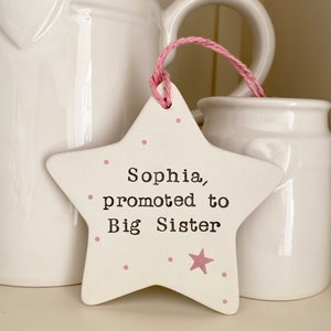 May include: White ceramic star with pink polka dots and a pink star. The star has the text "Sophia, promoted to Big Sister" written on it.