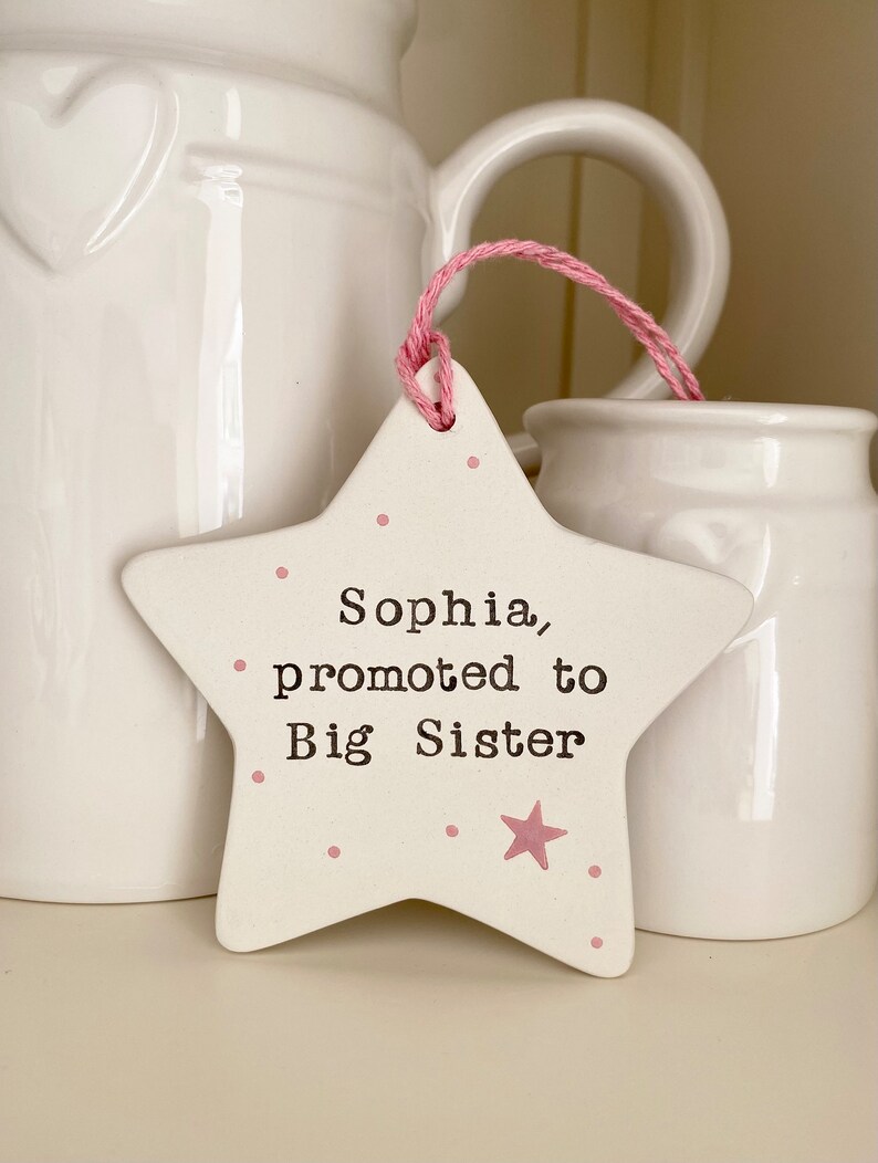 May include: White ceramic star with pink polka dots and a pink star. The star has the text "Sophia, promoted to Big Sister" written on it.