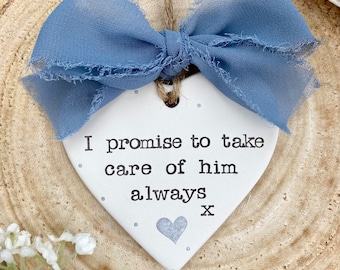 May include: A white heart-shaped ceramic ornament featuring the black printed text I promise to take care of him always x. The ornament is decorated with small light blue dots and a light blue heart, and it is topped with a frayed blue ribbon.
