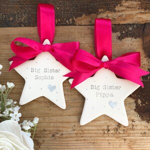 May include: Two white ceramic star decorations with pink ribbon bows. The stars have a silver glitter finish and the text "Big Sister Sophie" and "Big Sister Pippa" is written on them.