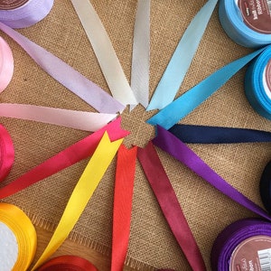 May include: A selection of satin ribbon spools in various colours, including lavender, light blue, yellow, red, and burgundy. The spools are arranged in a circle on a hessian background.