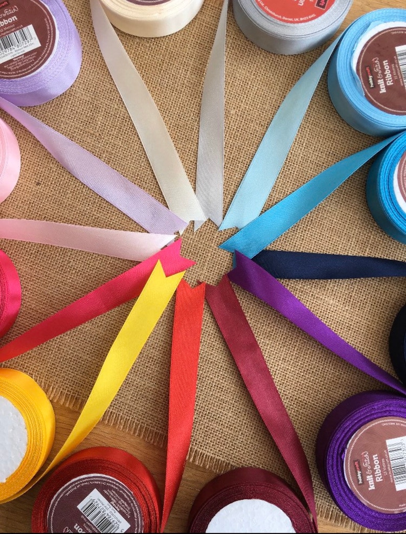 May include: A selection of satin ribbon spools in various colours, including lavender, light blue, yellow, red, and burgundy. The spools are arranged in a circle on a hessian background.
