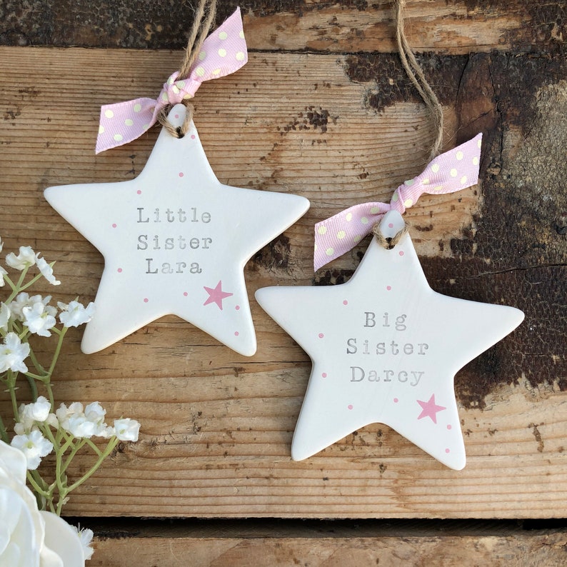 May include: Two white ceramic star ornaments with pink polka dots and pink text. The larger star says "Big Sister Darcy" and the smaller star says "Little Sister Lara".