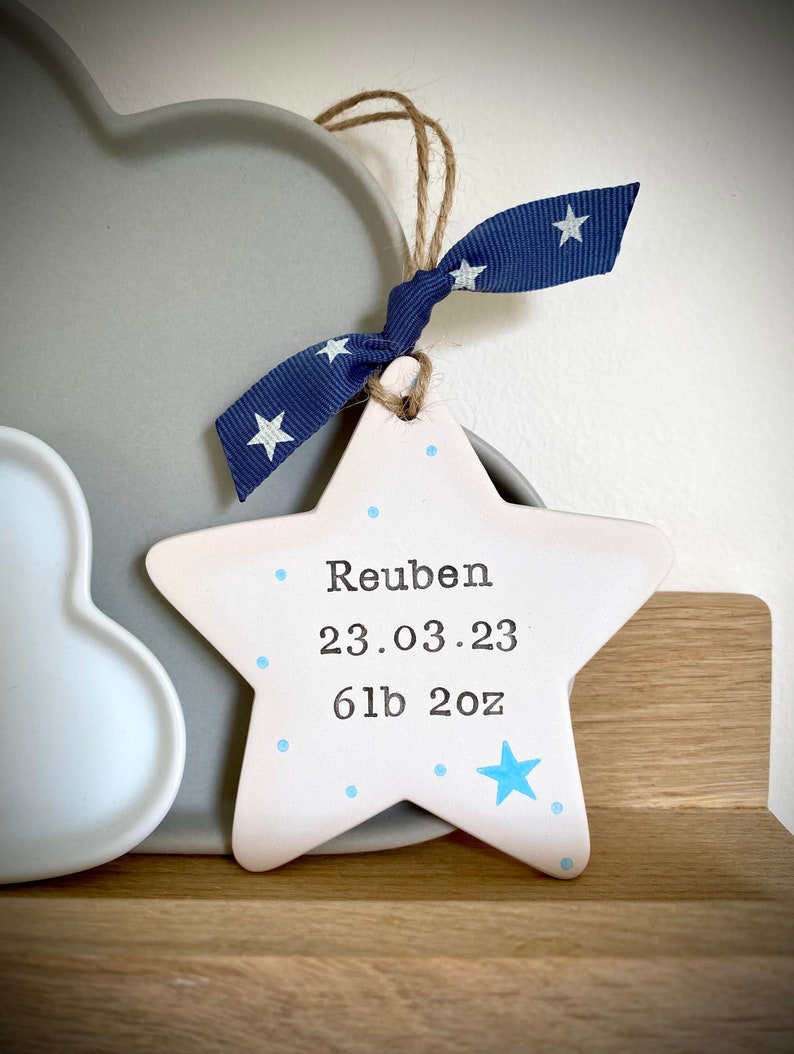 May include: A white ceramic star with blue dots and a blue star. The star has the name Reuben, the date 23.03.23, and the weight 6lb 2oz written on it.