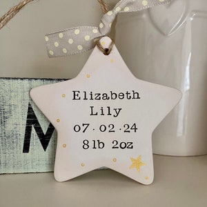May include: White ceramic star ornament with gold glitter accents. The star has the text "Elizabeth Lily 07.02.24 8lb 2oz" written on it.
