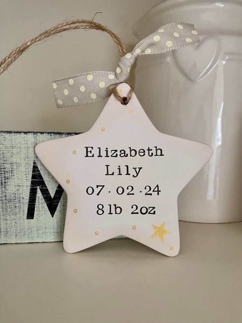 May include: White ceramic star ornament with gold glitter accents. The star has the text "Elizabeth Lily 07.02.24 8lb 2oz" written on it.