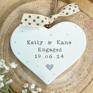 May include: White heart-shaped engagement ornament with the names "Kelly & Kane" and the date "19.06.24" printed in black. The ornament has a gray heart and polka dot ribbon. The ornament is displayed on a wooden surface.
