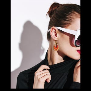 May include: A close-up of a person wearing a black turtleneck and white sunglasses with dark lenses. The person is wearing a gold teardrop earring with red and orange accents. The earring is hanging from the ear.