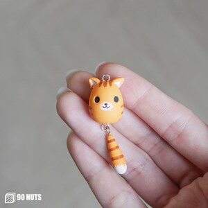 Può includere: Ciondolo a forma di gatto in argilla arancione e marrone con la punta della coda bianca. Il gatto ha un viso carino con occhi neri e un naso rosa.