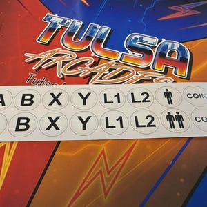 May include: A sticker sheet with white circular labels featuring black text and symbols. The labels include letters (A, B, X, Y, L1, L2), a coin symbol, and a restroom symbol. The background features the text "Tulsa Arcades".